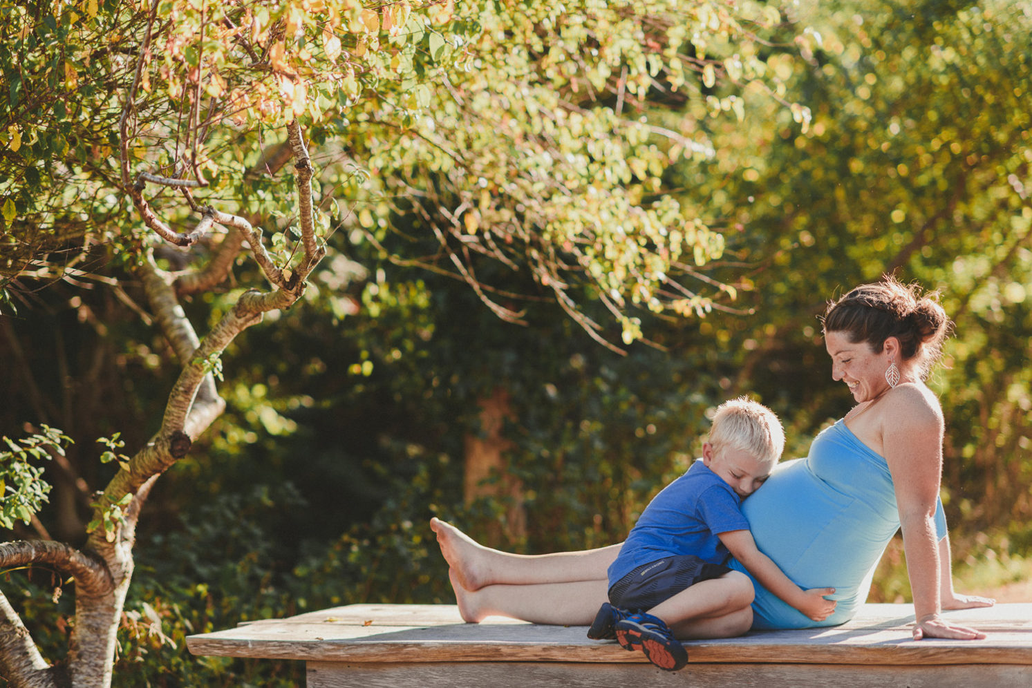 young pregnant woman sitting on a picnic bench with her young son hugging her pregnant belly-hornby island lifestyle maternity session