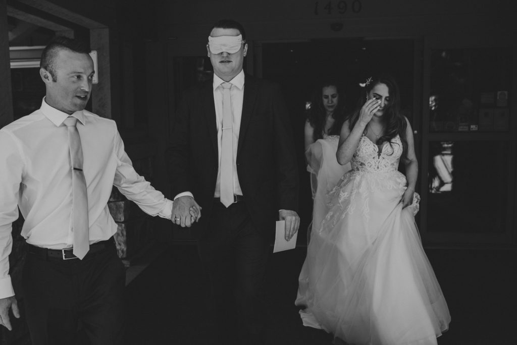 best man leading blindfolded groom out of a hotel  lobby followed by the bride