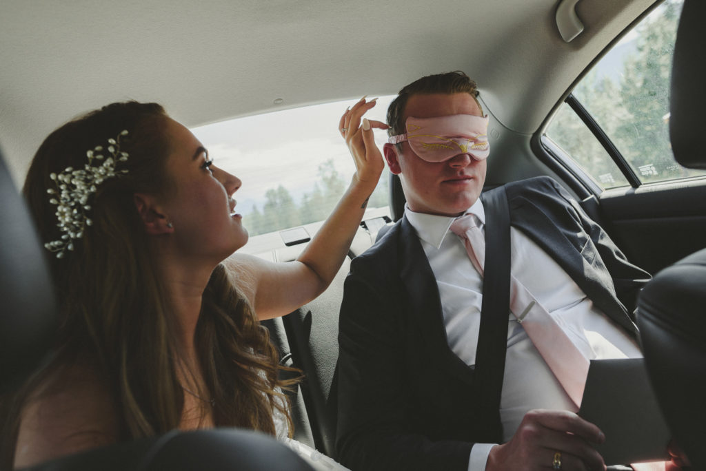 bride fixing her blindfolded groom's hair in the backseat of a car
