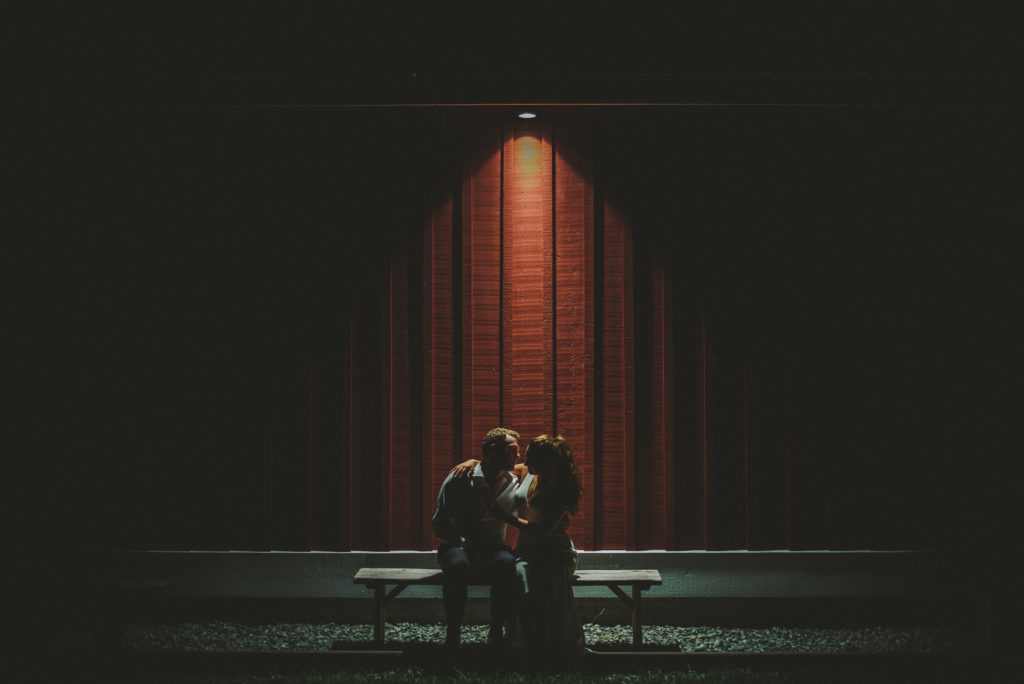 portrait of bride & groom under a light against a red barn wall late at night
