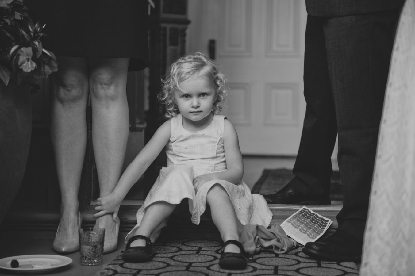 young girl in a dress sitting amongst adult legs holding her mother's ankle at a wedding