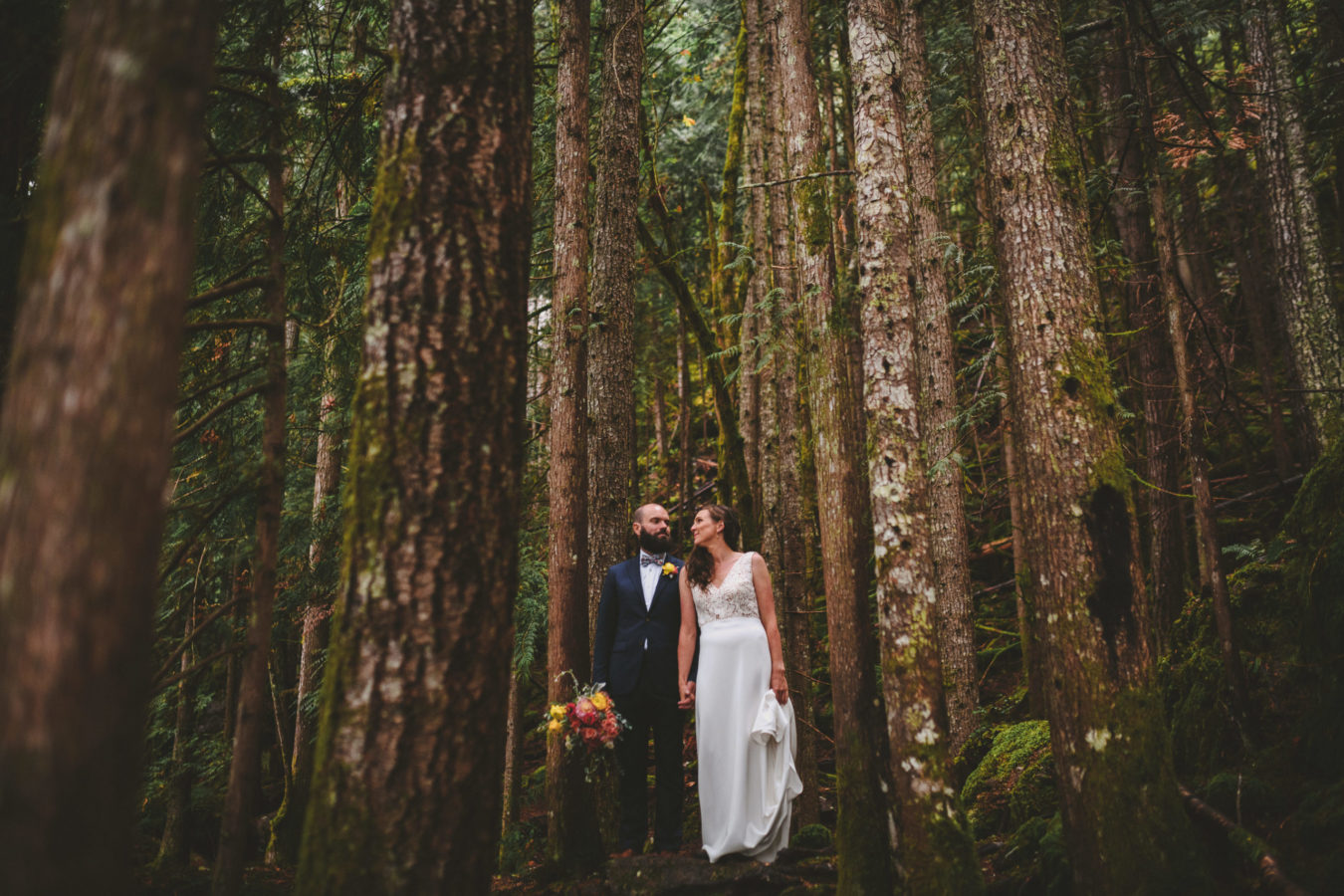 bride and groom holding hands and looking at each other amongst tall trees in the forest