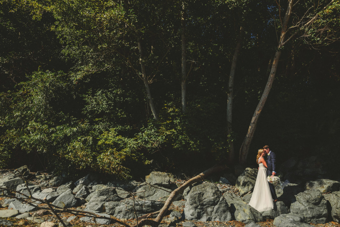 bride and groom kissing at the edge of a beach with tall trees behind them