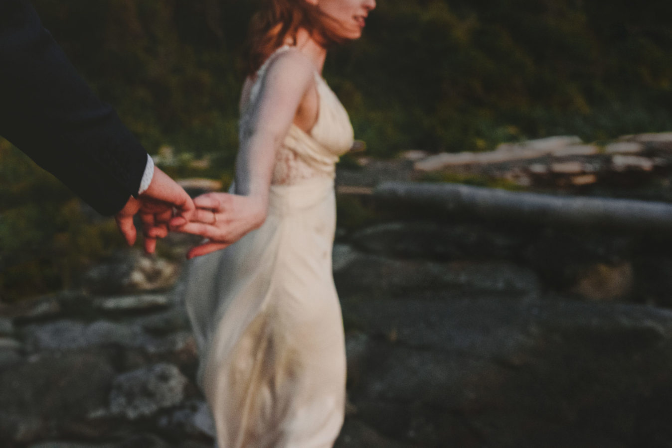 out of focus photo of a bride reaching back and holding her groom's hand walking along a beach after sunset