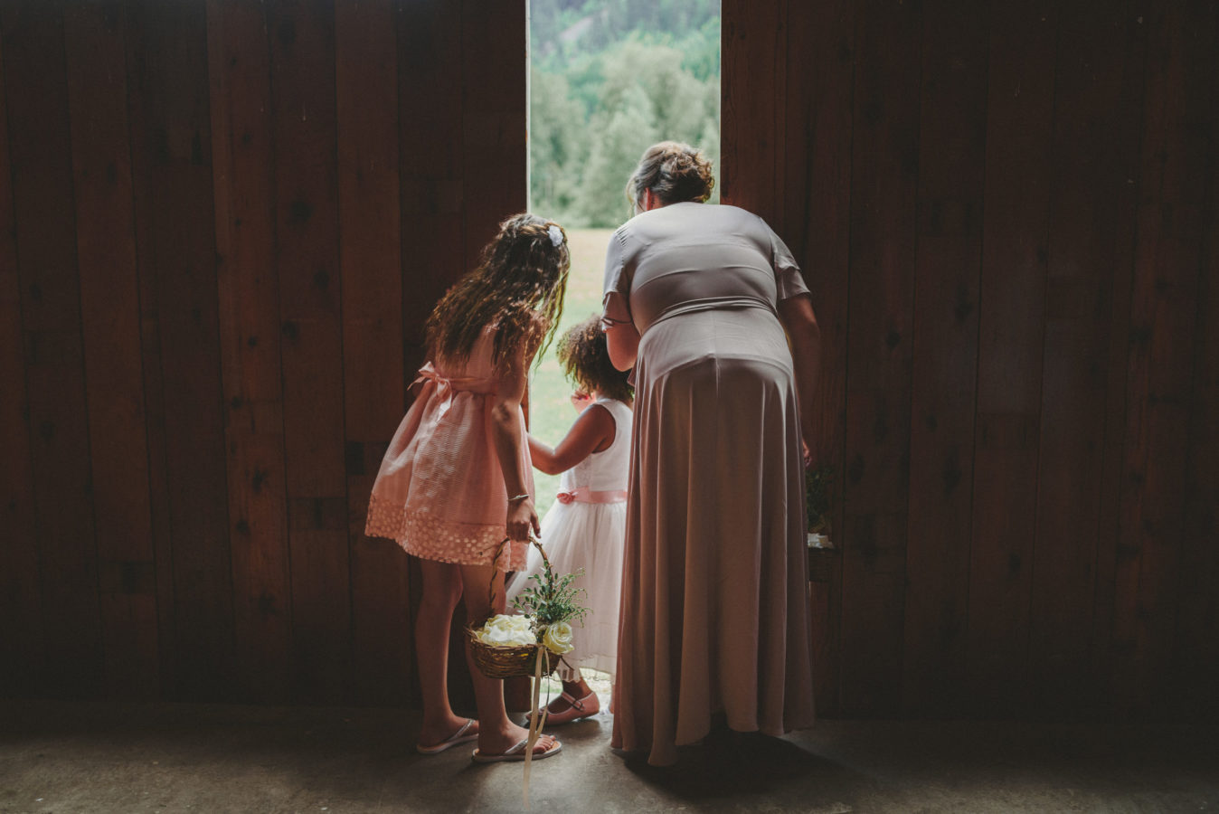two flower girls and a bridesmaid peeking out of a barn door waiting to walk down the aisle