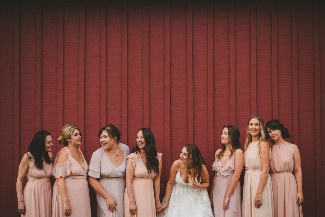 bride and bridesmaids laughing while leaning up against the red wall of a large barn