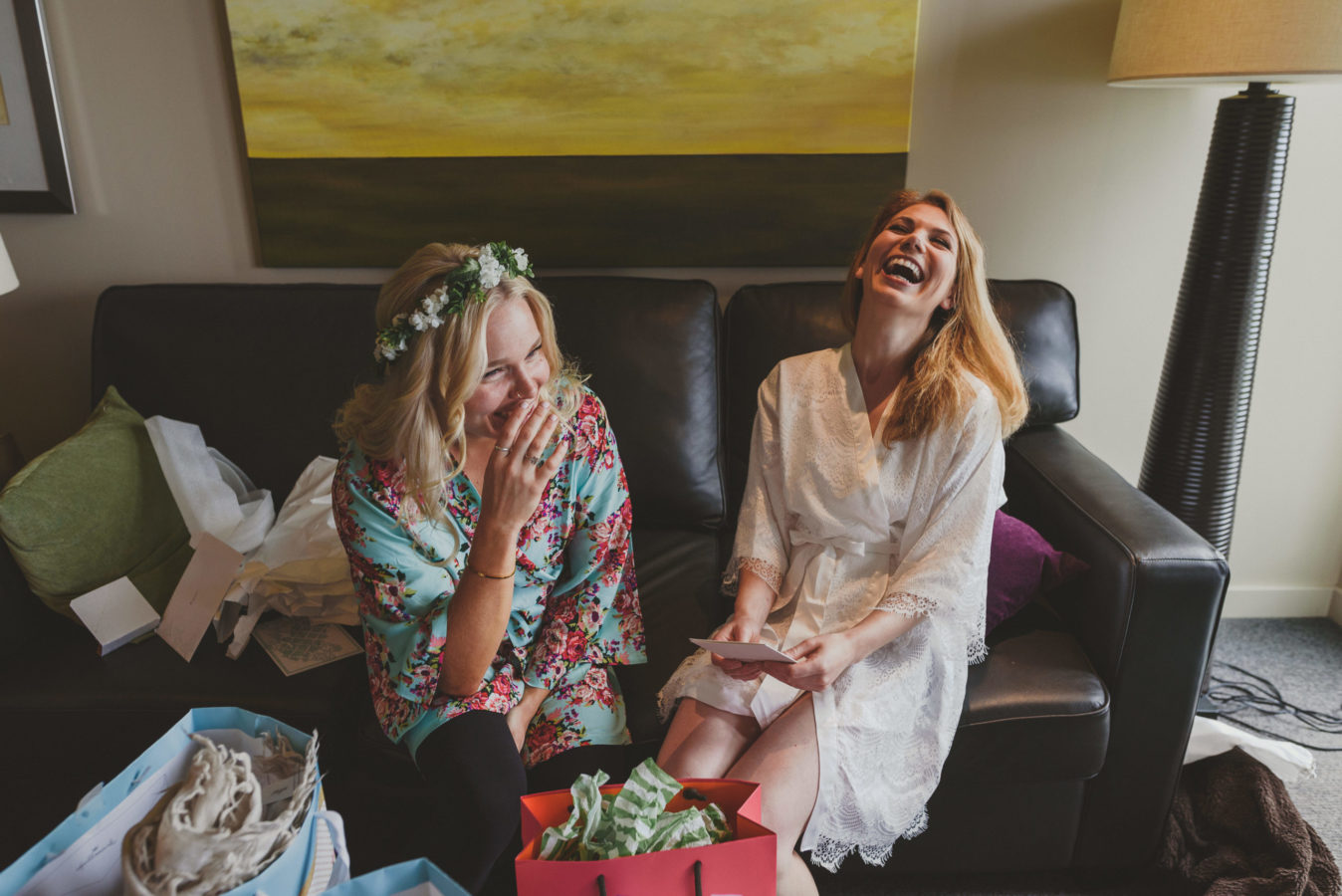 bride and bridesmaid in house coats laughing on a couch while getting ready