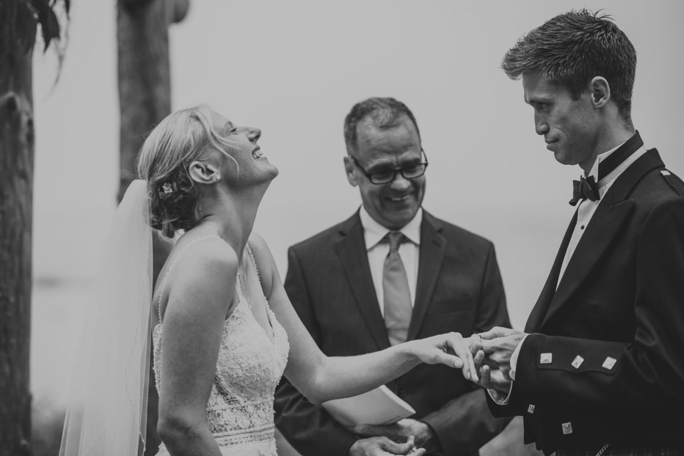 bride throwing her head back laughing while her groom tries to put on her ring during their wedding ceremony