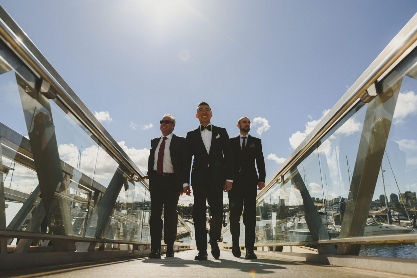groom and two groomsmen walking up a dock with the sun & blue sky behind them