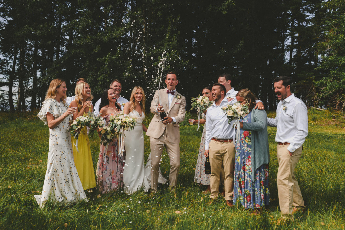 groom popping a bottle of champagne surrounded by his bride and wedding party all laughing & surprised