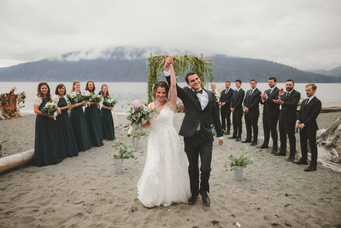 bride and groom walking back up the aisle after getting married on the beach with their hands held high with their wedding party in the background
