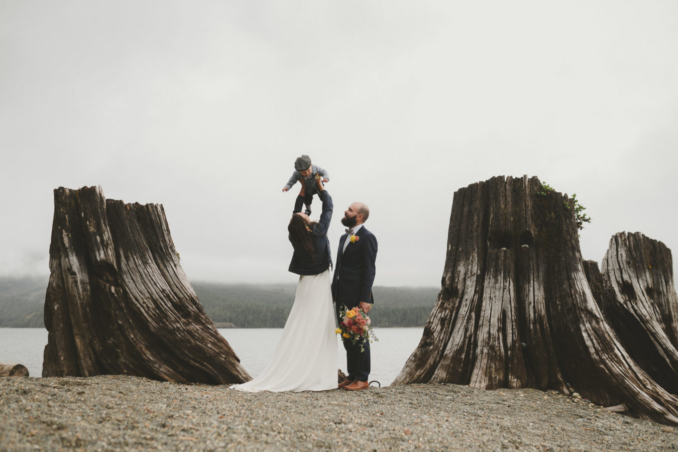 bride and groom on the beach by two huge stumps while bride gently tosses their toddler in the air above her