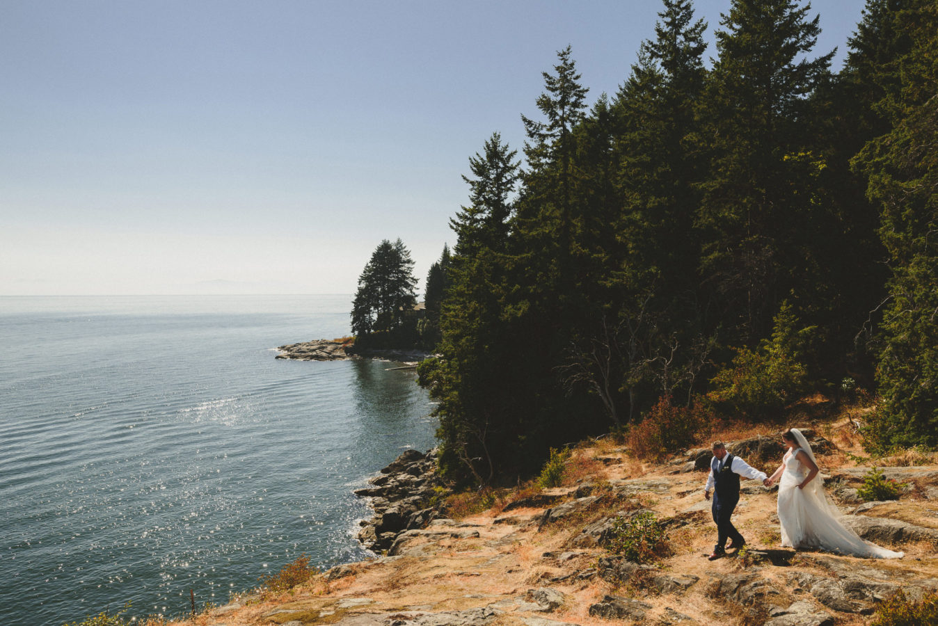 looking down on a bride and groom walking hand in hand out along a bluff overlooking the ocean