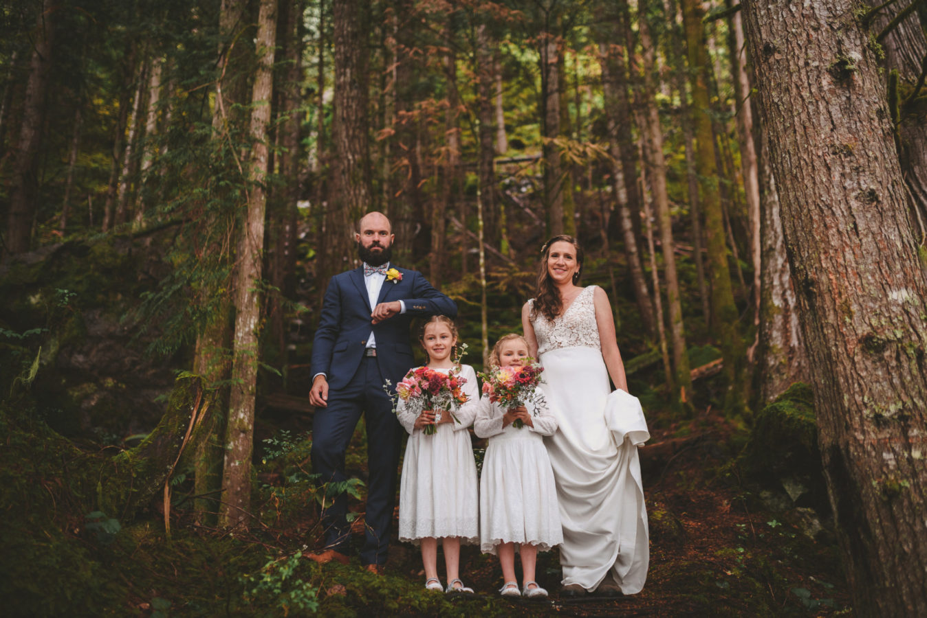 bride, groom and two flower girls standing among tall trees in the forest for a portrait