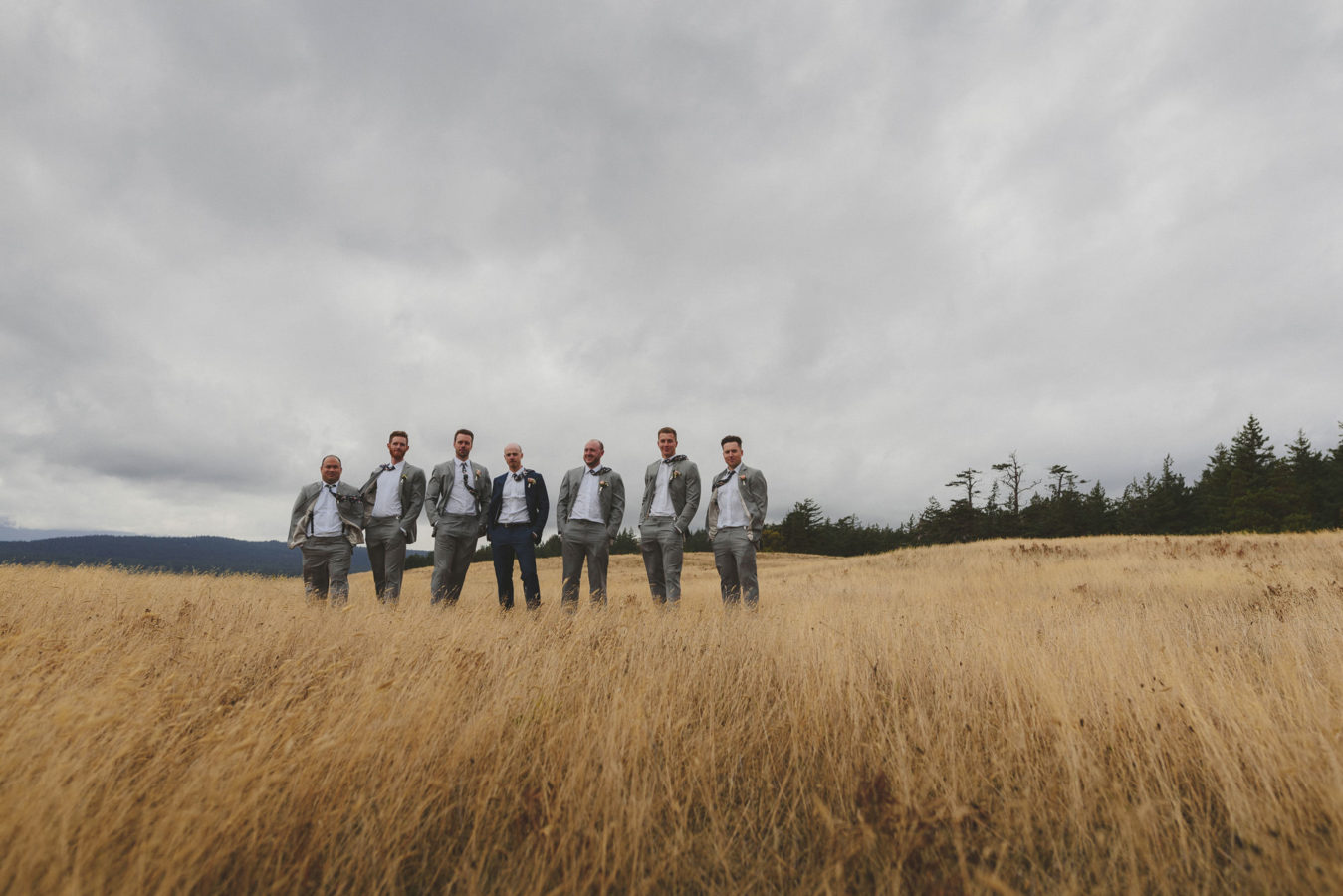 looking across a wind swept grassy field at a groom and size groomsmen standing for a portrait in the wind with a stormy sky above