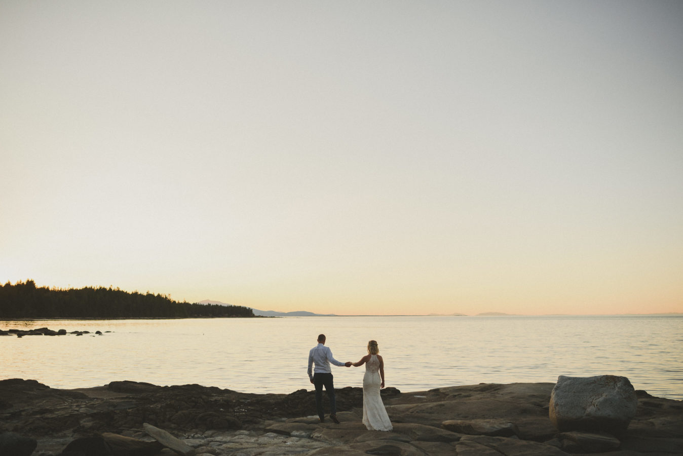 bride and groom holding hands and walking towards the ocean on a rocky shore at sunset