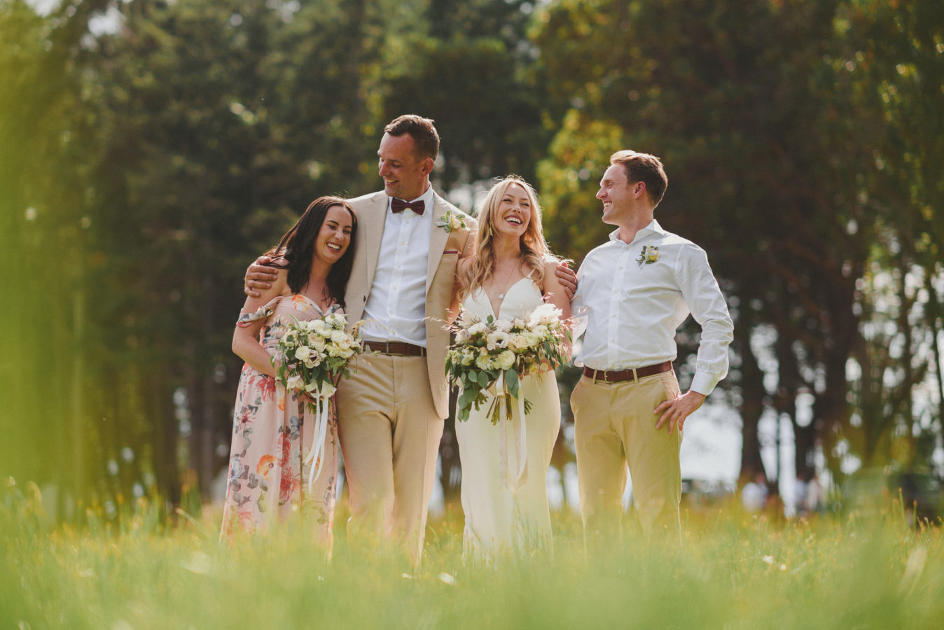 bride, groom and their maid-of-honour and best-man standing and laughing in a field of tall grass with the forest behind them