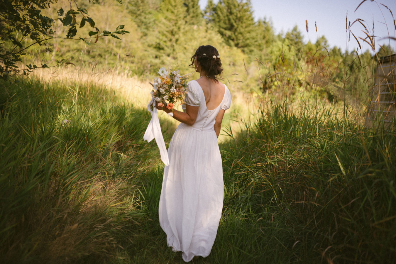 back of a bride in her wedding dress walking away in a field holding her bouquet of flowers