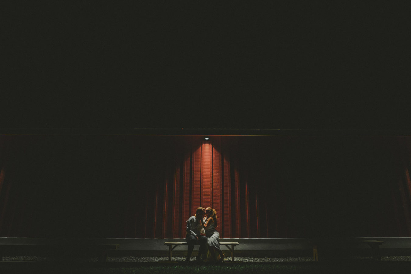 bride and groom at night on a bench next to a red barn with one light shining down upon them