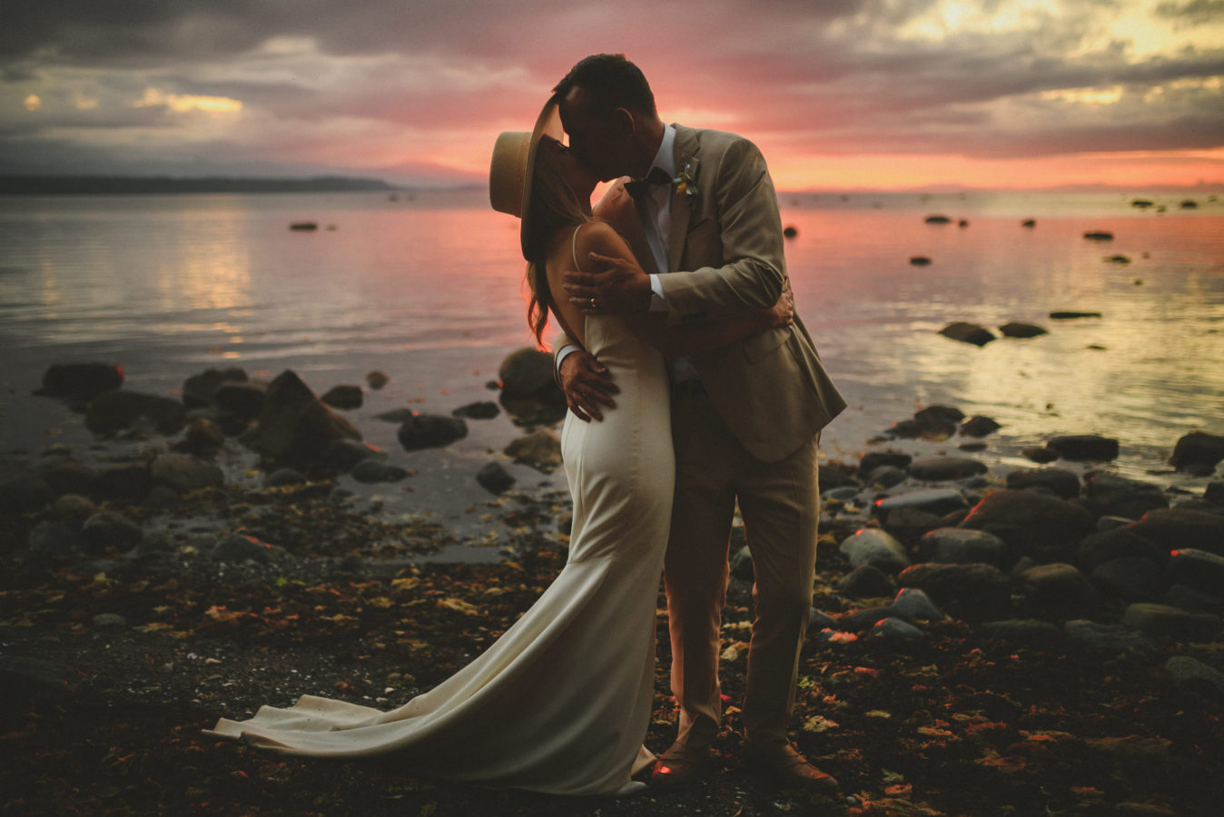 bride and groom kissing in a deep embrace on the edge of a rocky beach at sunset