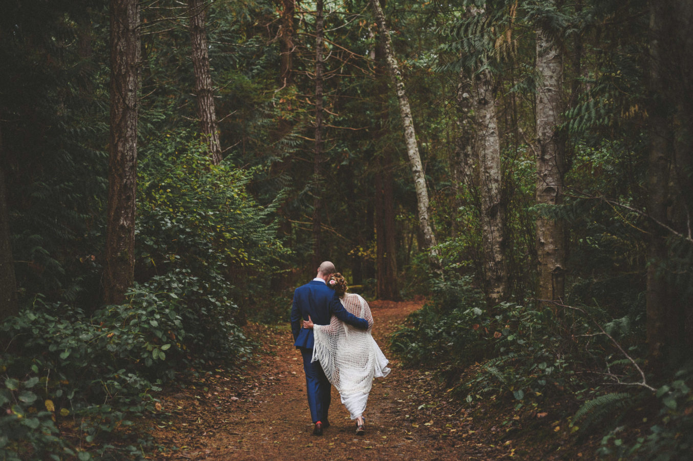 bride and groom walking arm in arm down a forest path surrounded by tall trees