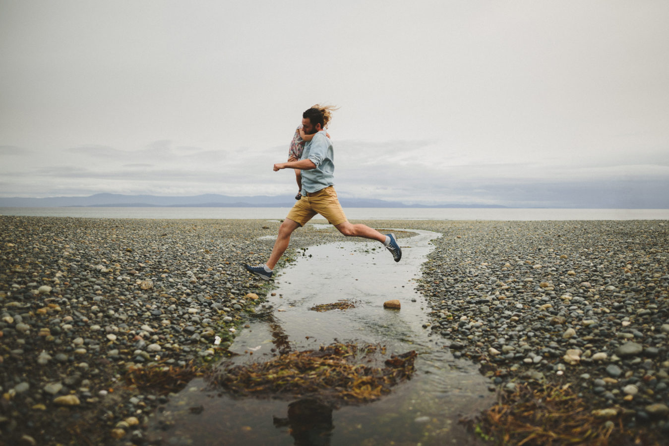 dad holding a kid jumping over a stream running down the beach