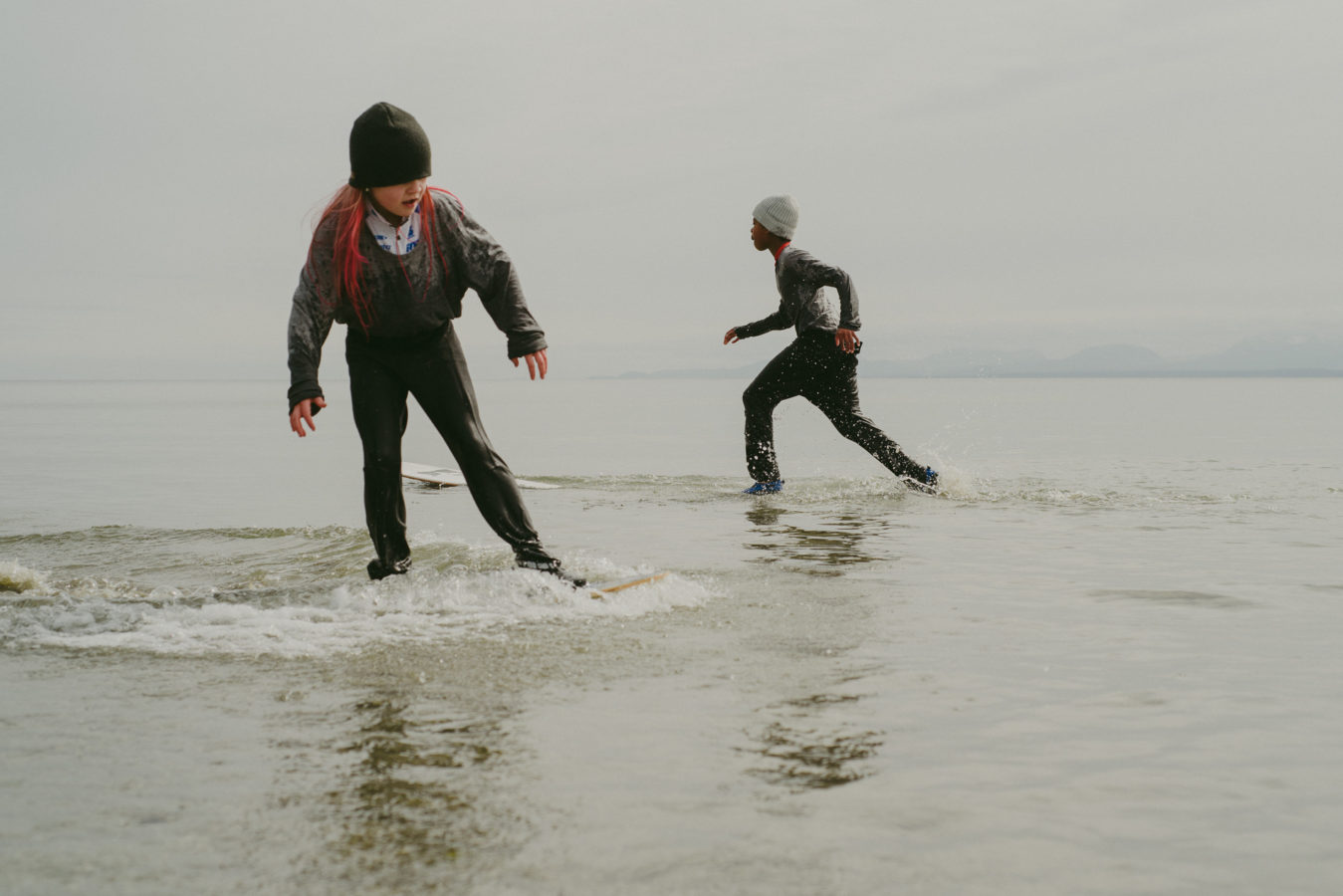 two kids skim boarding in winter clothes and toques