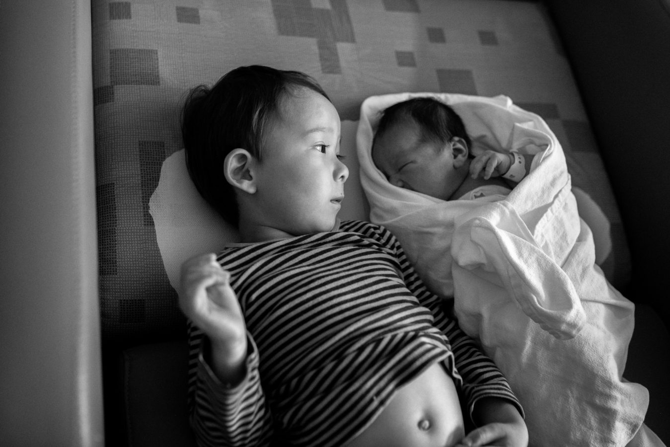 young boy lying down looking at his new baby swaddled