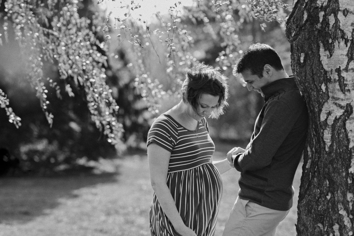 pregnant woman and her husband standing under a backlit willow tree