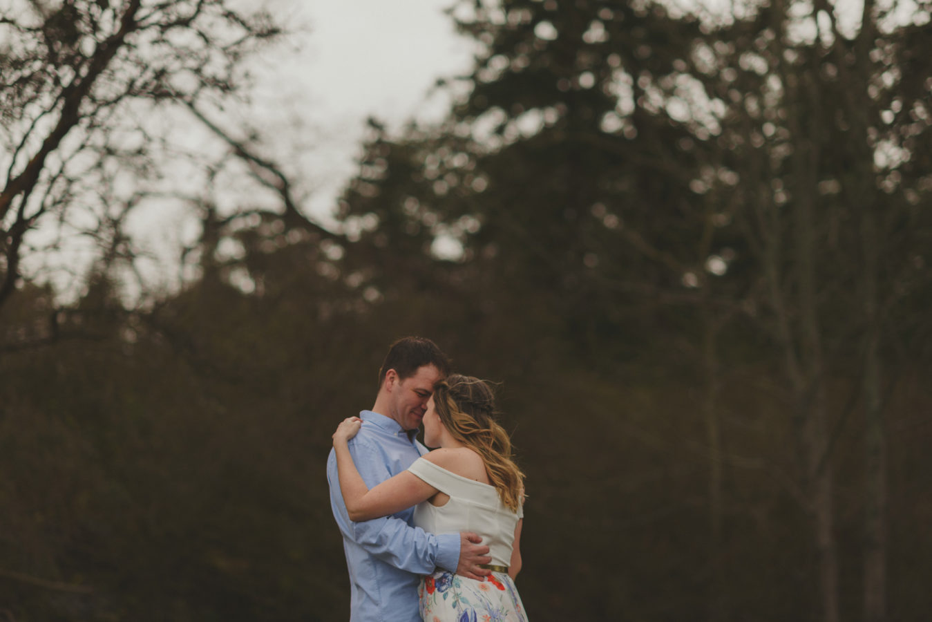 engaged couple snuggling against the dark trees