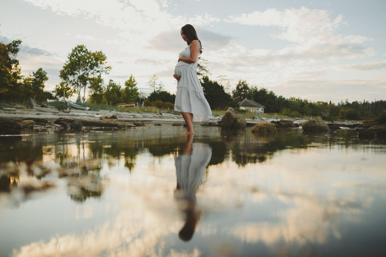 pregnant woman holding her belly standing in the water at the beach edge with her & the sunset reflected in the water