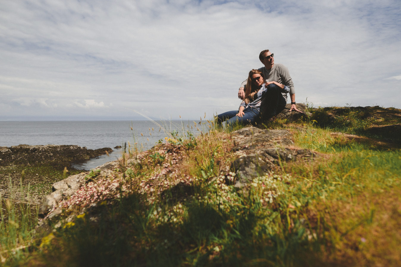 man and woman sitting and snuggling on a rocky bluff overlooking the ocean