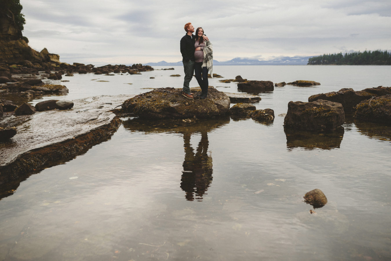 pregnant woman and her man embracing on the sandstone rocks reflected in the still ocean