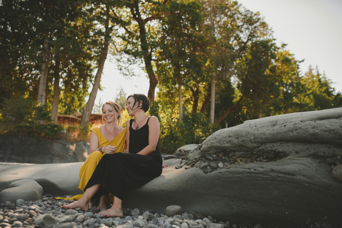 two women sitting close together and laughing on large rocks on the beach with the sunset light behind them