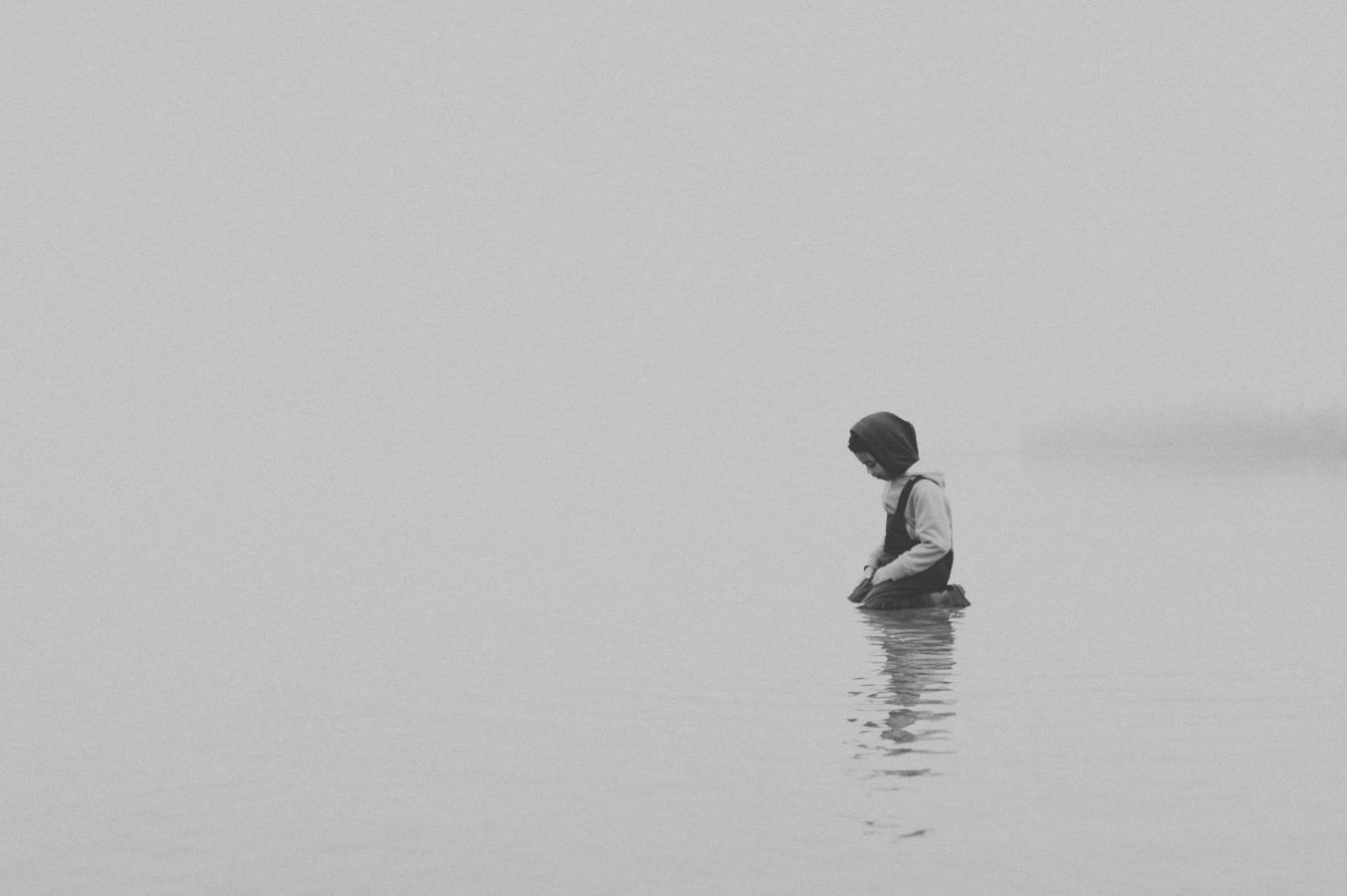 a lone child kneels in the calm ocean wearing a toque and rain pants in the fog