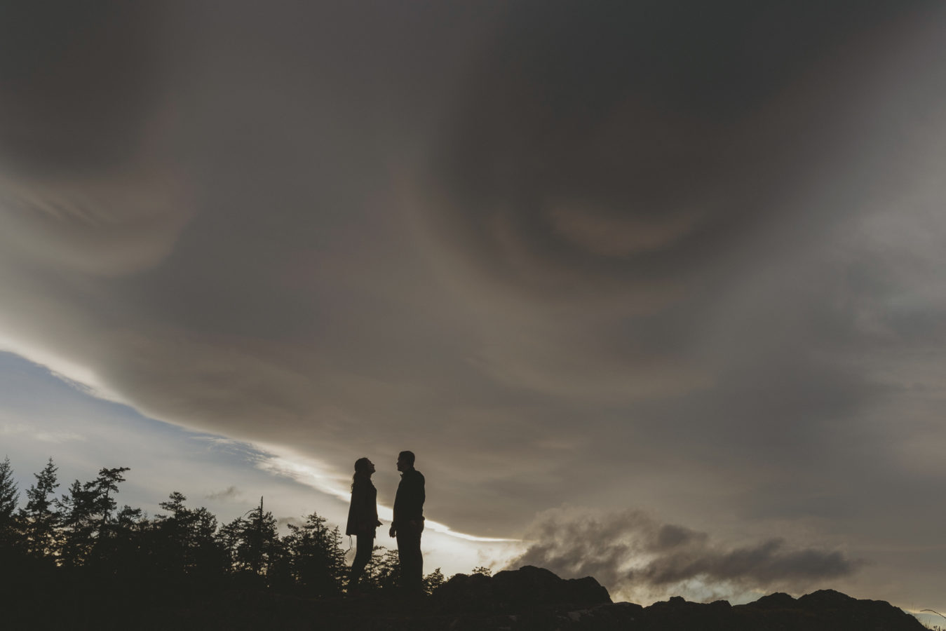man and woman standing on a rocky bluff with a dark dramatic sky behind them