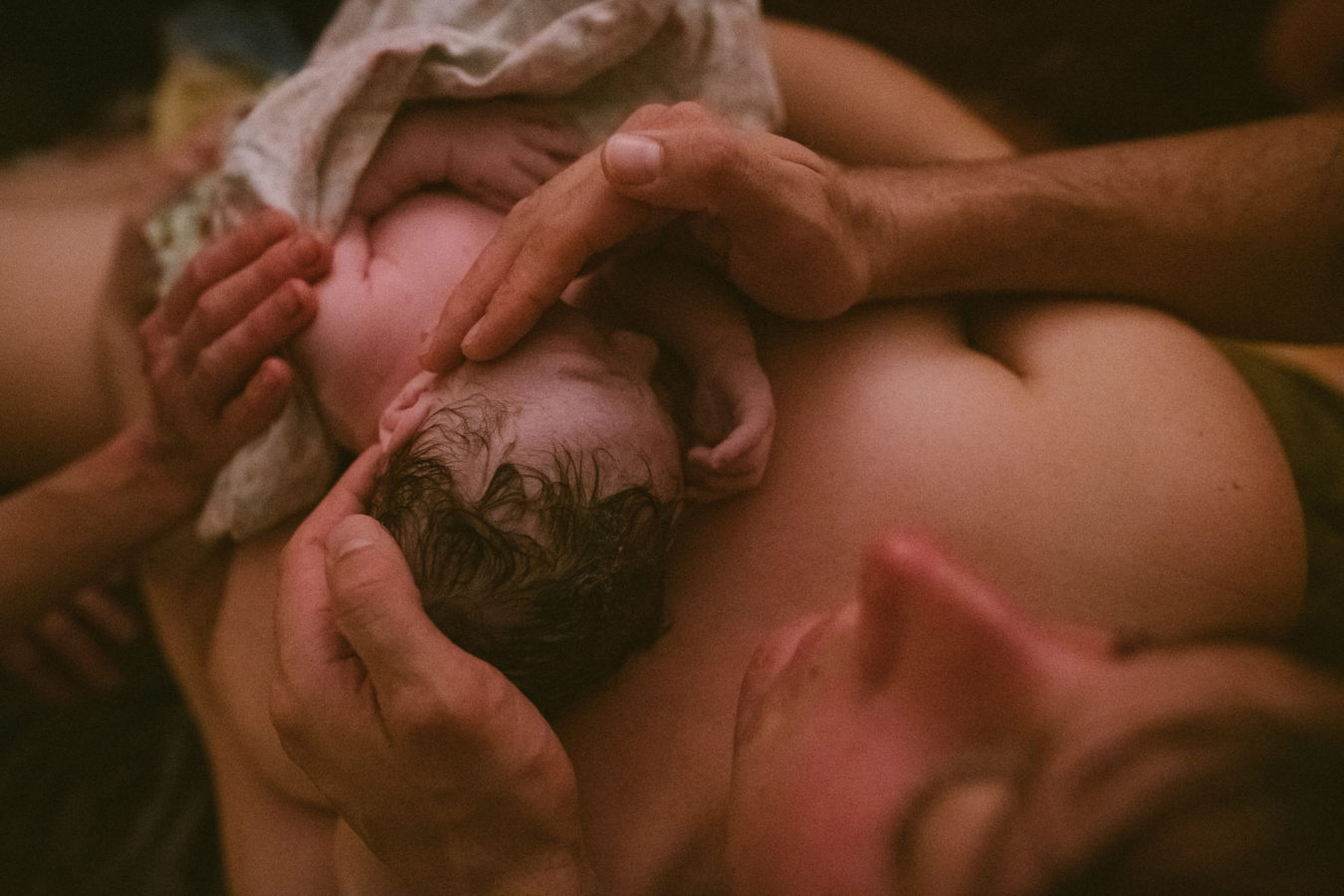 just born baby lying on his mother's chest with his father's hands shielding him from the light of the room