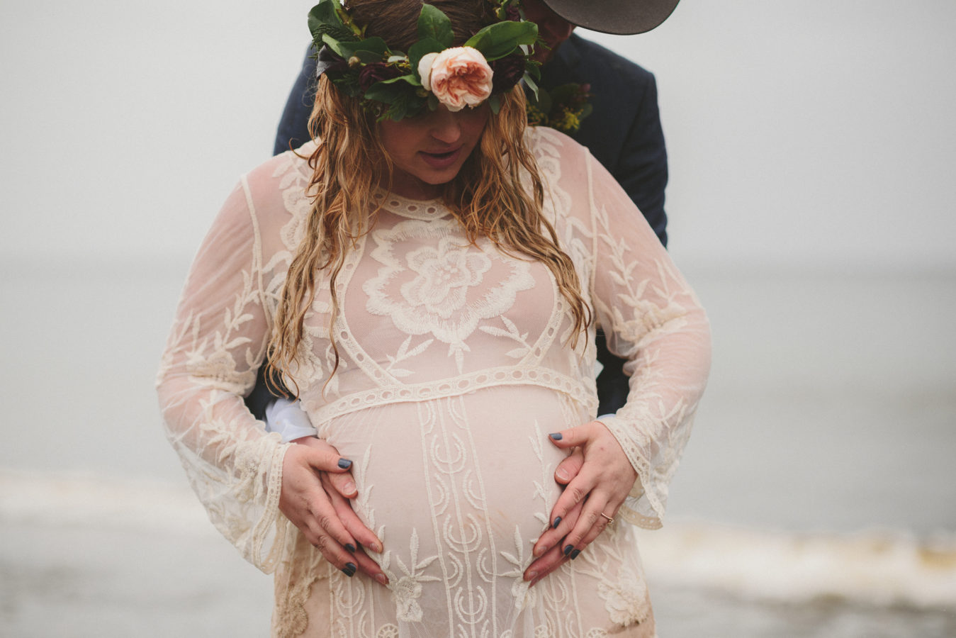 groom standing behind his pregnant bride holding her belly as they're both soaked with rain