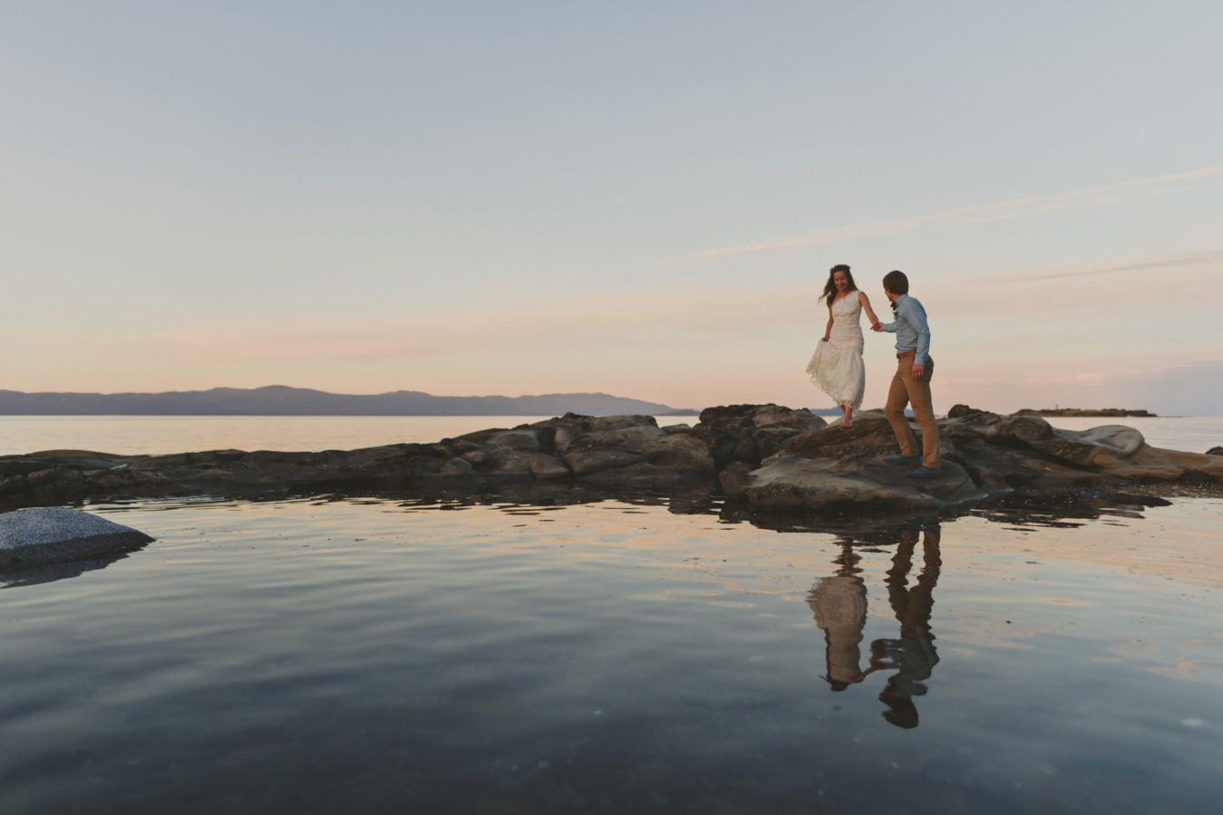 bride and groom walking along sandstone rocks in the water at sunset