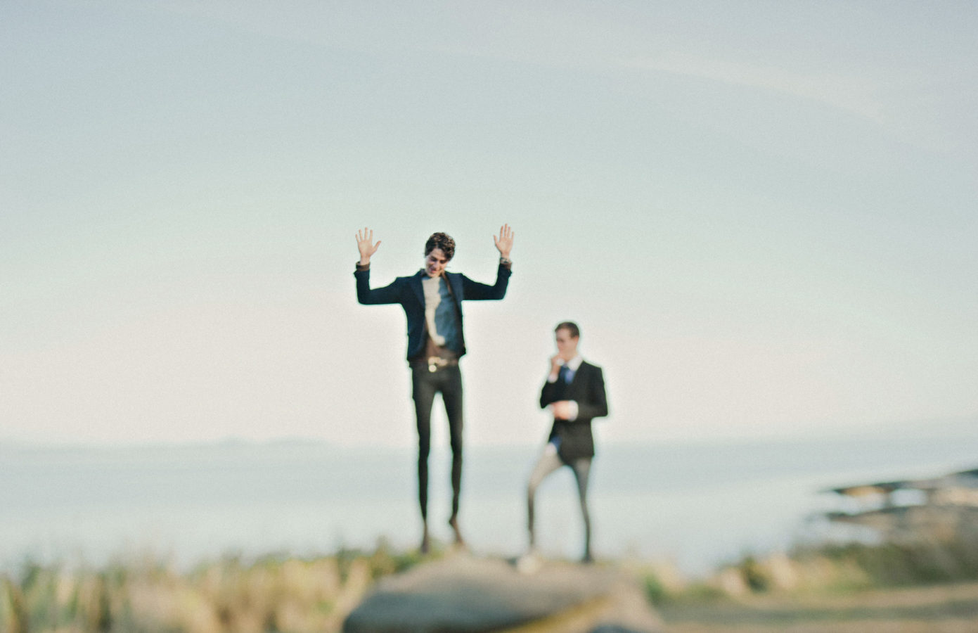 two grooms on a bluff by the ocean with one of them jumping in the air