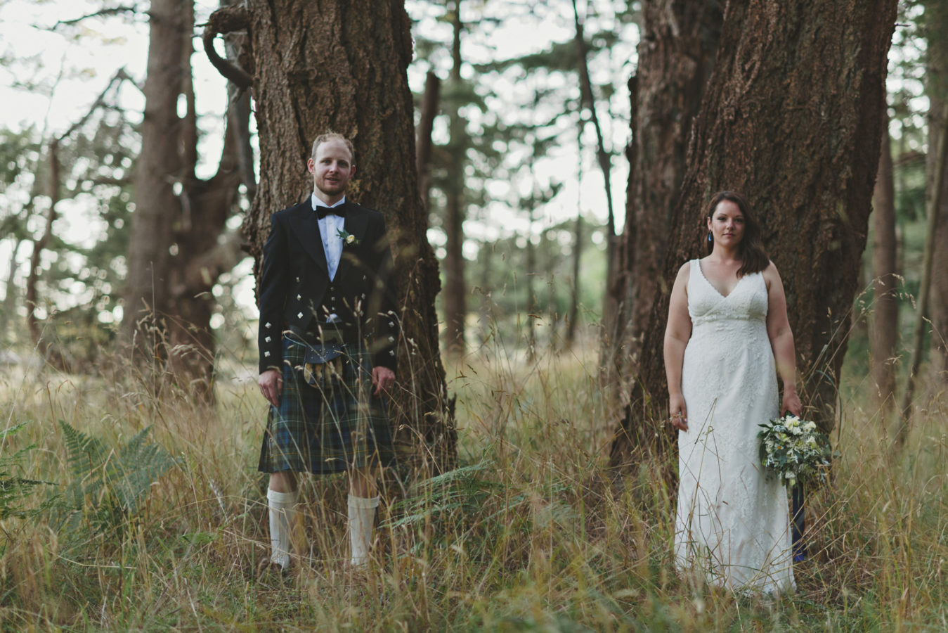 bride and groom each standing against an old growth tree looking deadpan into the camera