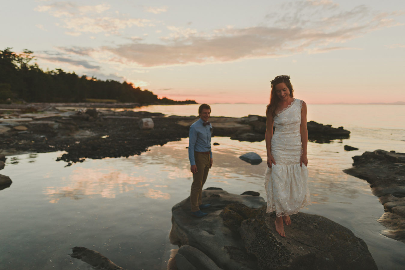 groom looking at bride as she walks along the sandstone rocks at the ocean's edge surrounded by the sunset