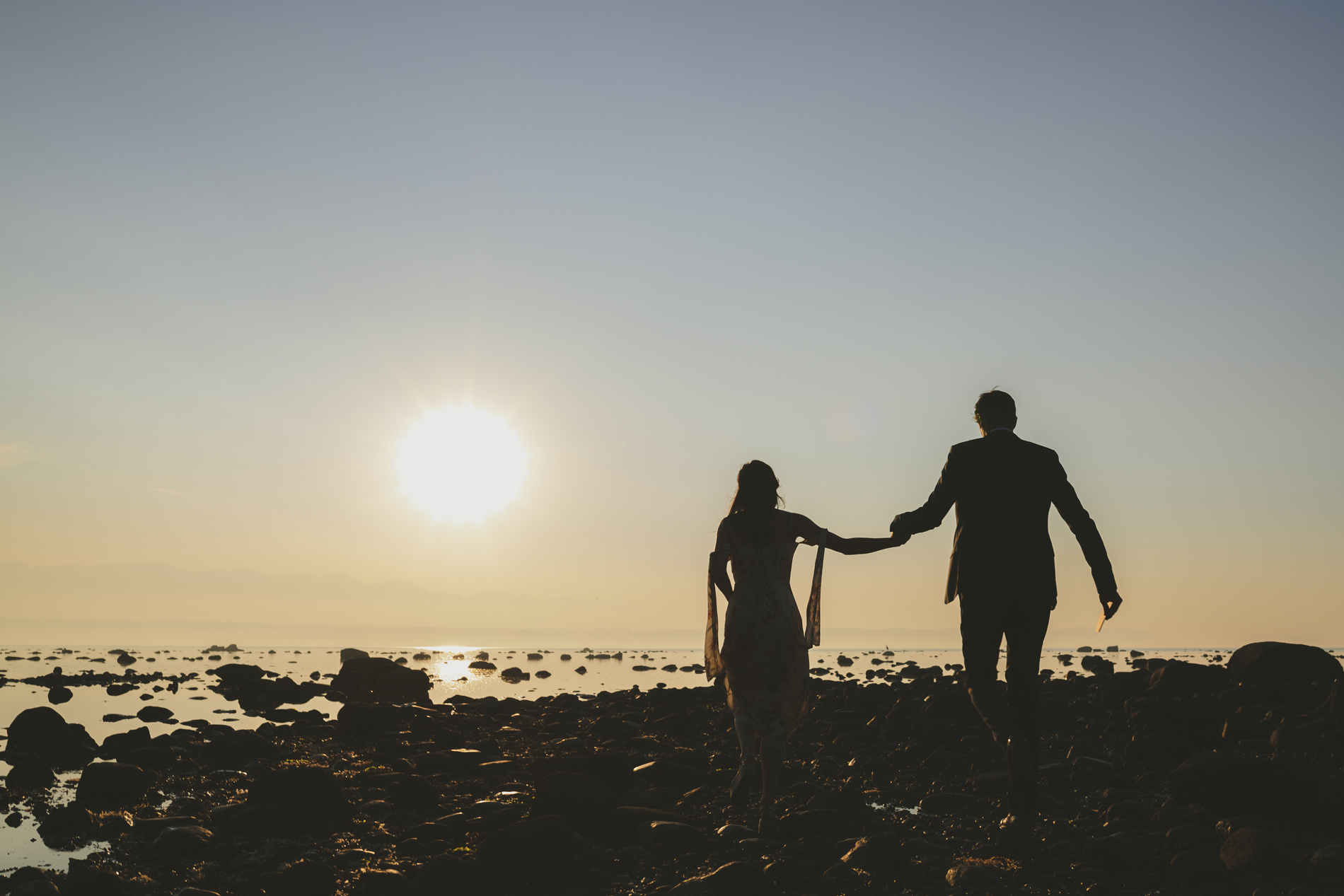 couple holding hands walking along a rocky beach silhouted by the sunset