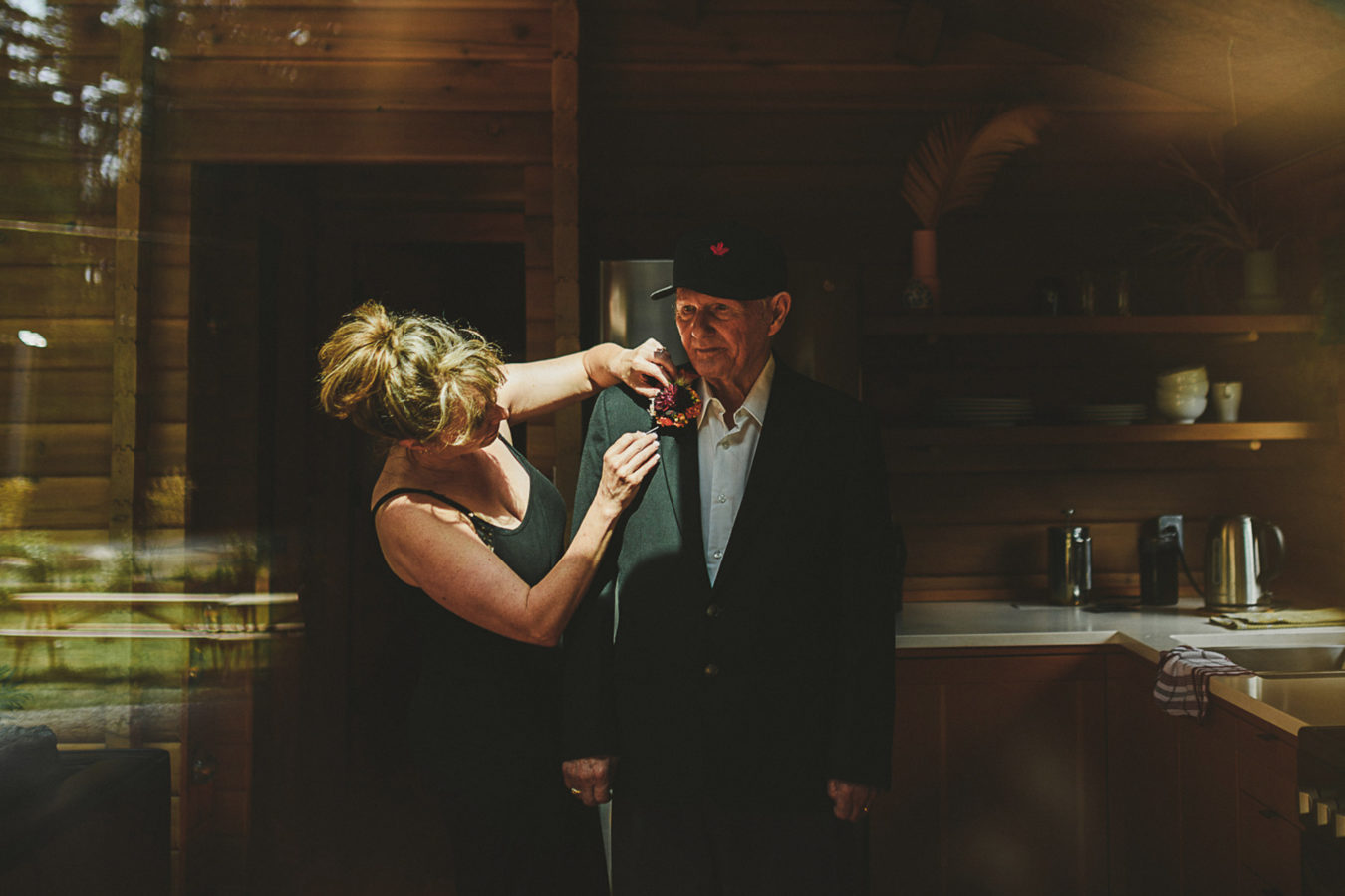 looking into a window at a woman pinning a boutonierre on her elderly father in a rustic cabin