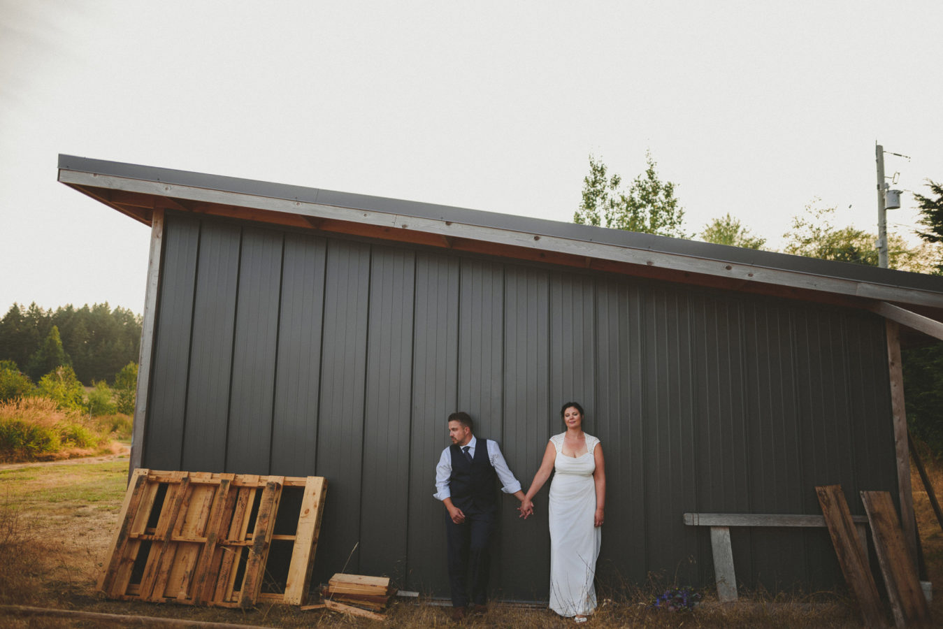 bride and groom leaning against a metal sided out building in a field on their wedding day