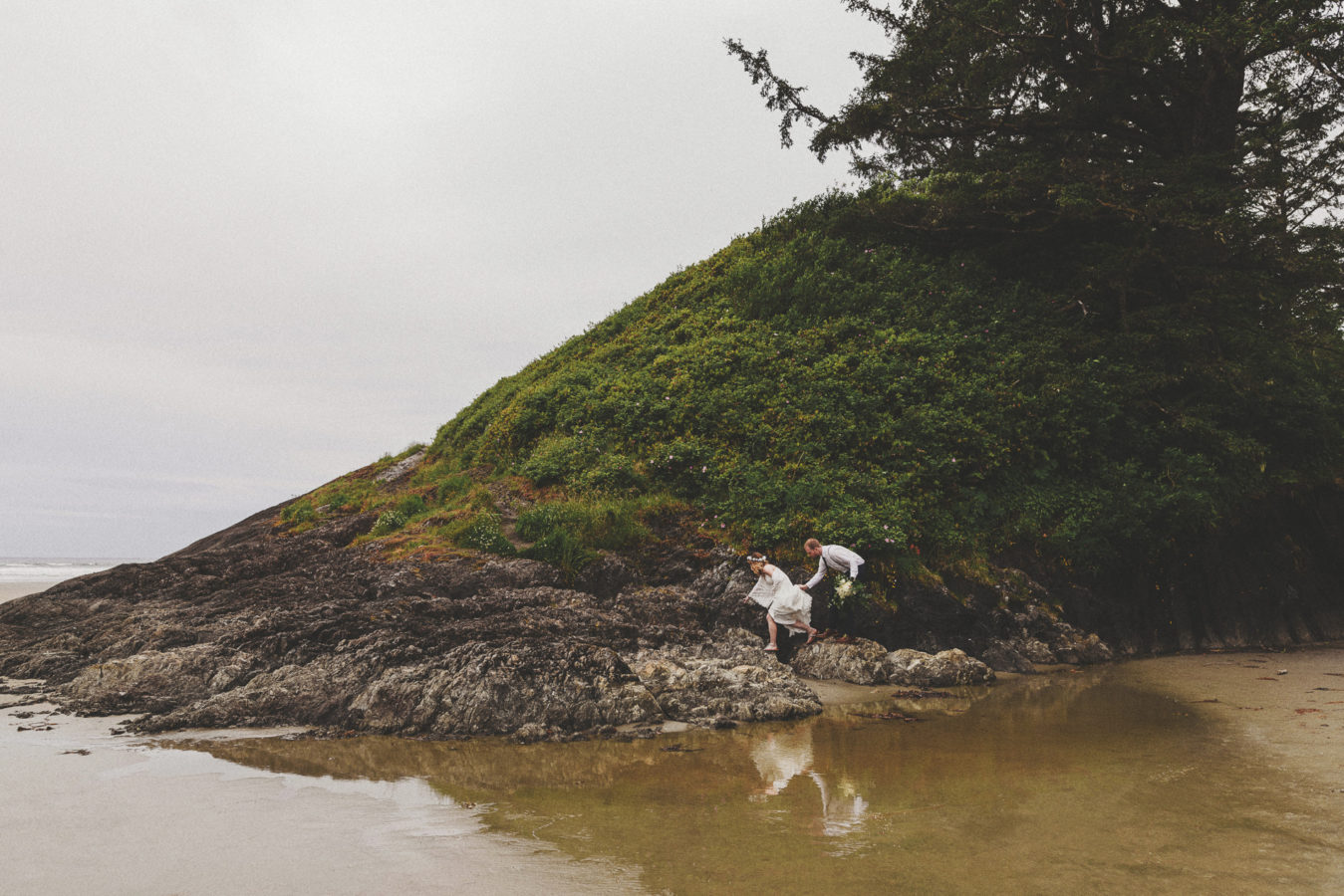 bride and groom clambering on a rocky bluff at the ocean's edge with their refection in the still water below them