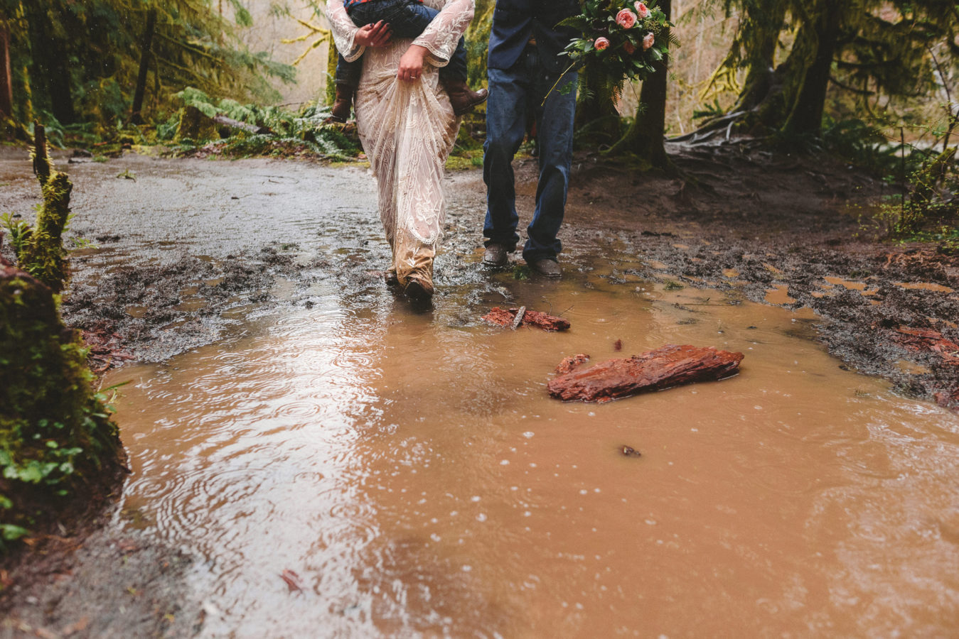 bride and groom walking together into a big mud puddle on a path in the forest on their wedding day while she holds their young child in her arms