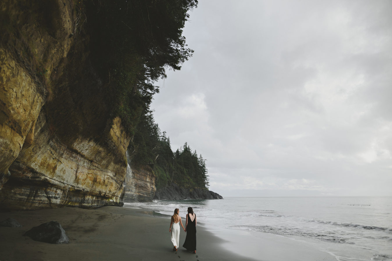 two brides walking down the edge of a beach towards a waterfall at the cliff's edge